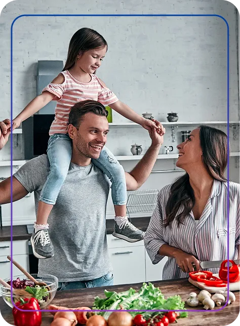 Man carrying smiling young girl on his shoulders while a woman prepares vegetables in a kitchen.