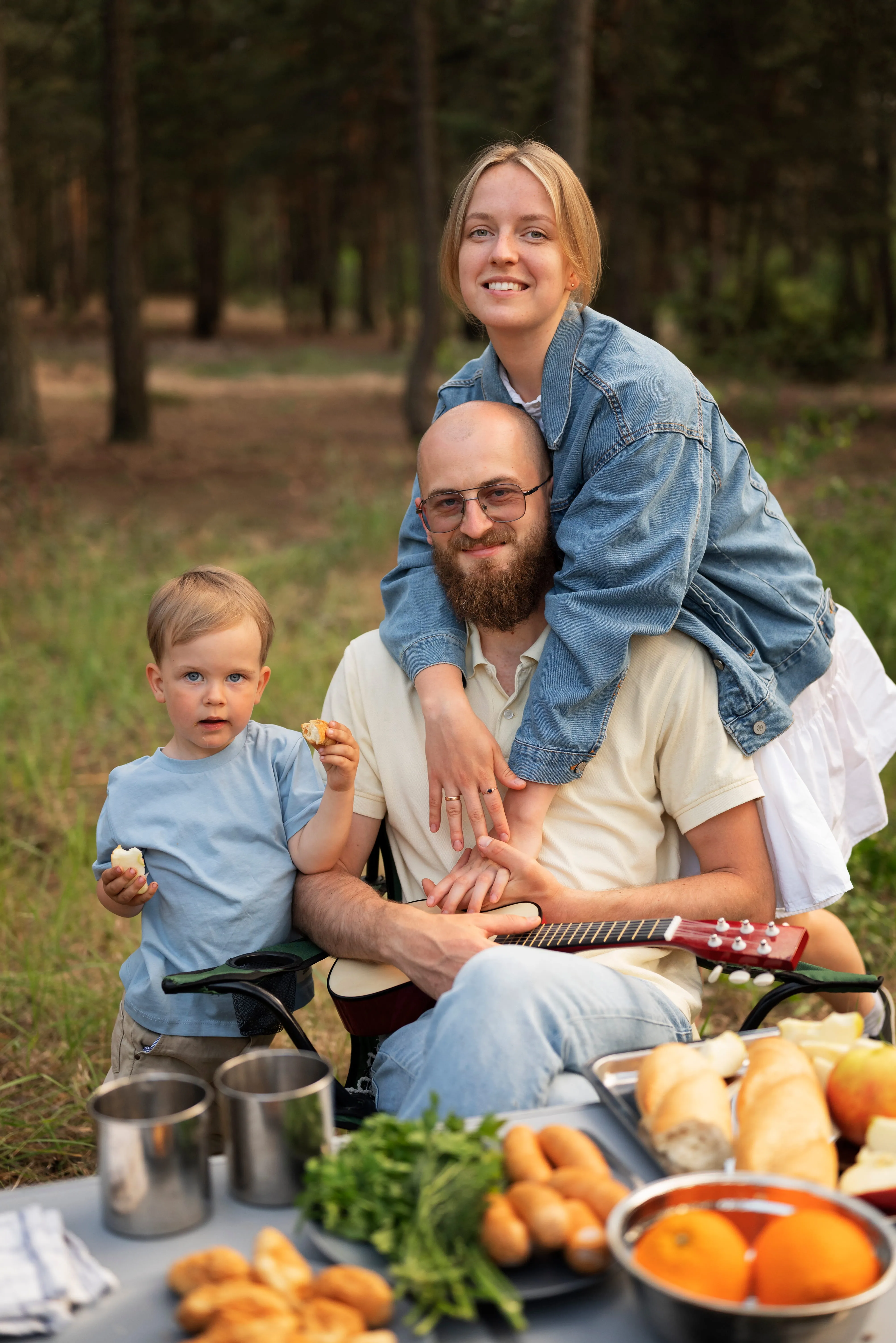 Smiling family outdoors with a man holding a guitar, a woman hugging him from behind, and a child eating bread, surrounded by picnic food.