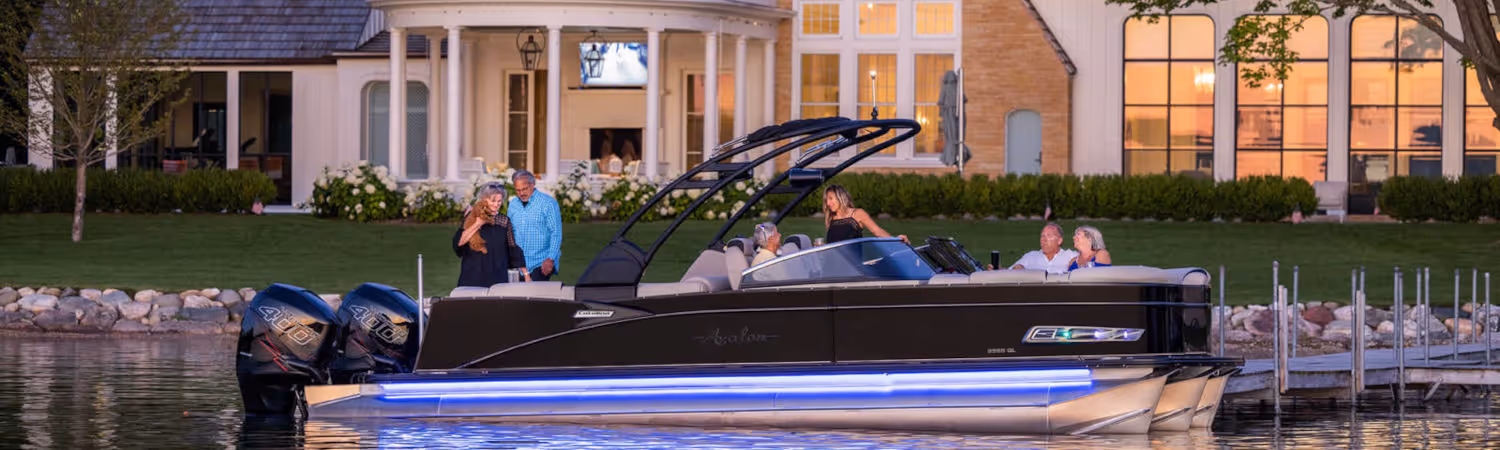 Six people enjoying a black Avalon pontoon boat with blue LED lights on a lake in front of a large house at sunset.