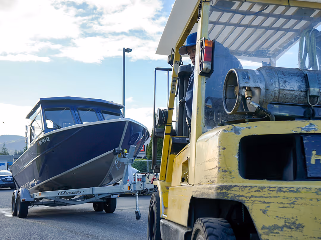 Yellow forklift towing a trailer with a blue boat on an asphalt surface.