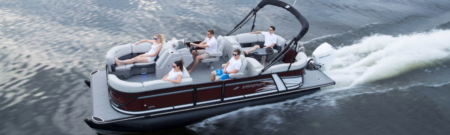 Pontoon boat cruising on water with five casually dressed people seated and one person steering.
