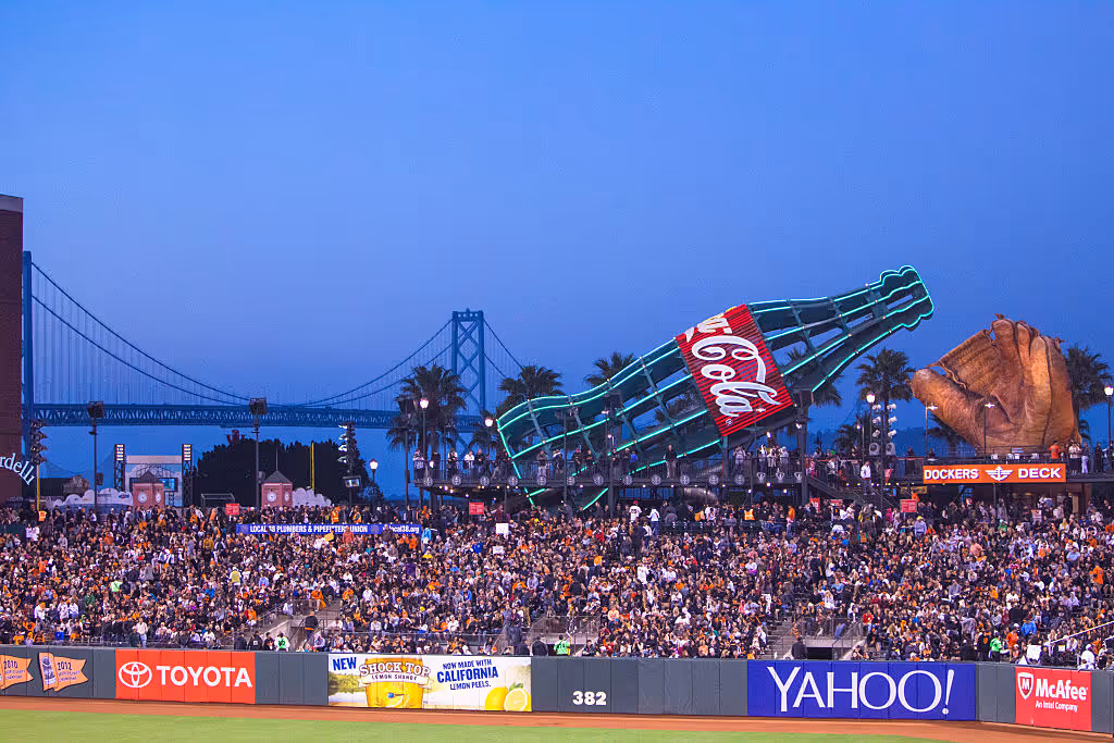 A packed baseball stadium at dusk with a large illuminated Coca-Cola bottle and oversized glove sculpture beyond the outfield, with a bridge visible in the background.