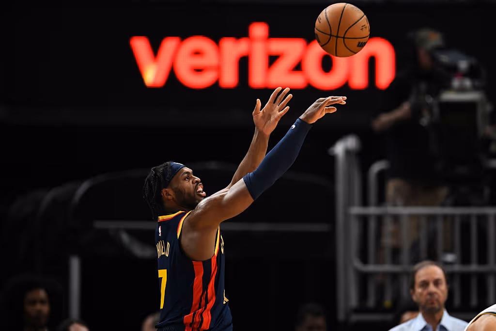 A basketball player in a dark uniform releases a shot mid-air, with a bright Verizon sign glowing behind him.