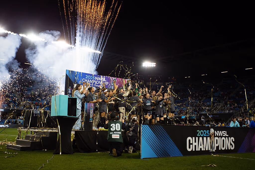 Women’s soccer team celebrating the 2025 NWSL Championship on a stage with the trophy raised, fireworks and gold streamers exploding above them in a packed stadium.
