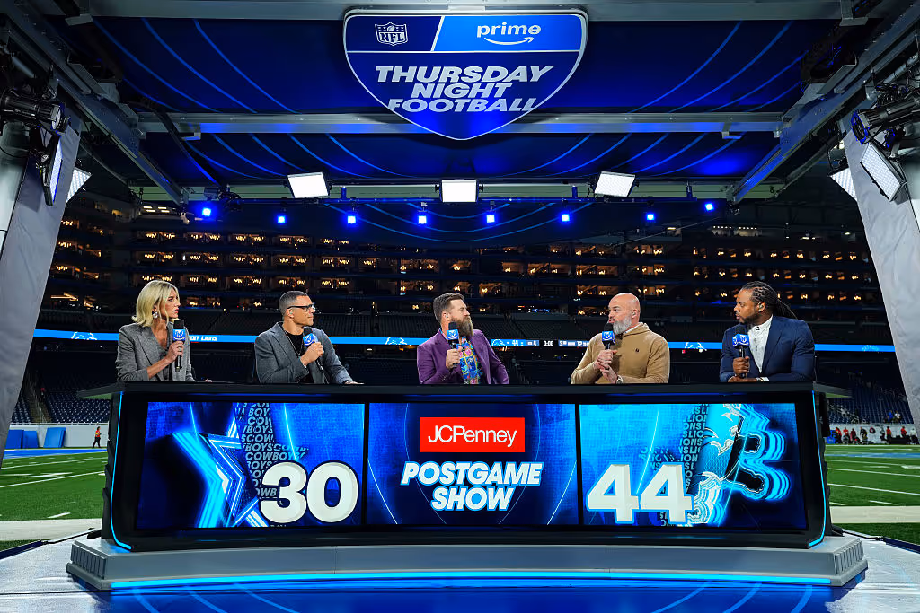 Broadcast analysts sit at a Thursday Night Football postgame desk on the field, discussing the game with stadium lights glowing behind them.