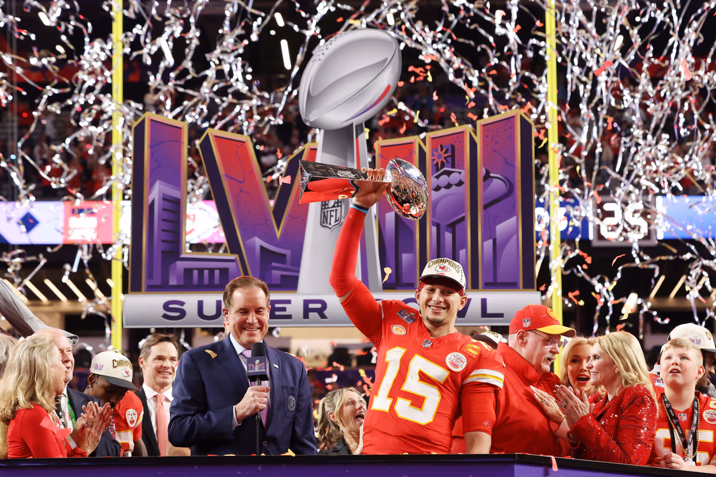 Image from a Super Bowl celebration showing a football player holding the championship trophy while broadcasters stand nearby.
