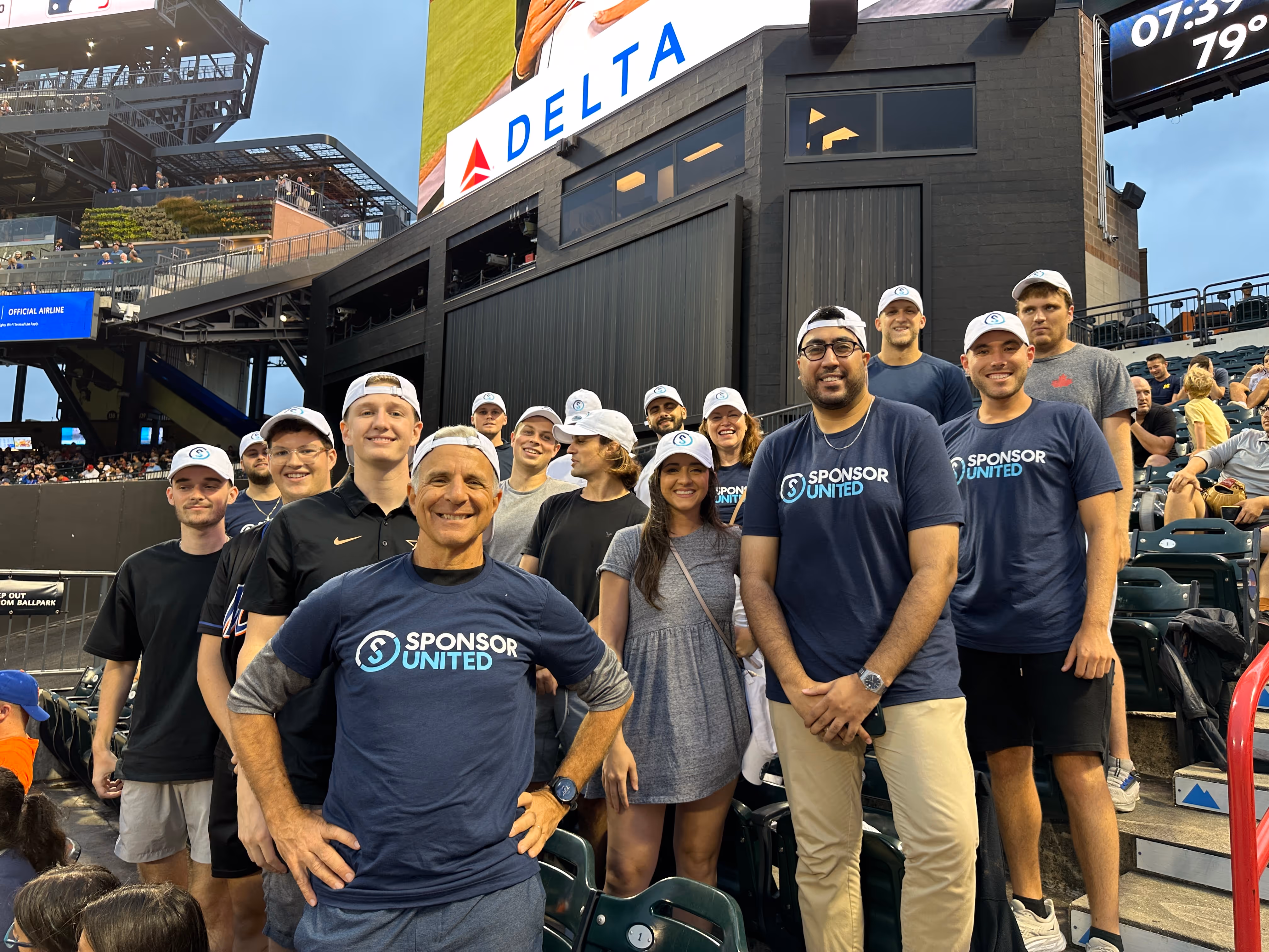 A group of SponsorUnited team members pose together in the stands at a baseball stadium, wearing matching shirts and hats during a company outing.