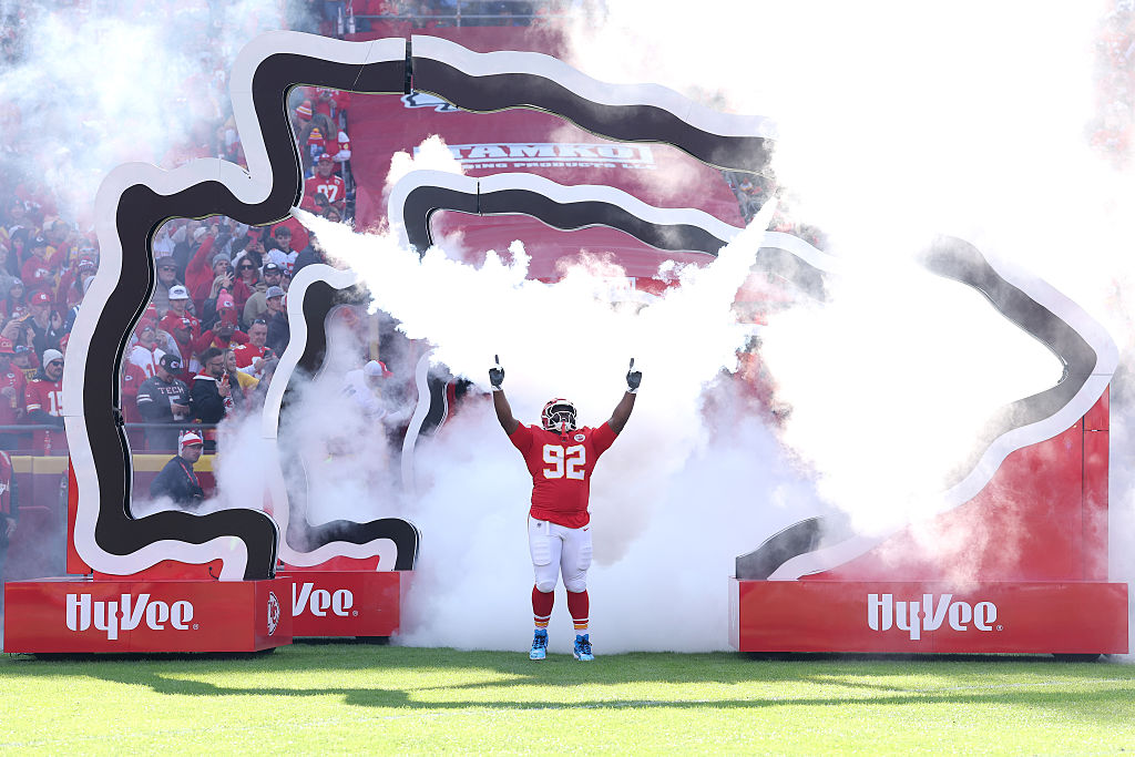 A Kansas City Chiefs player emerges through smoke with arms raised during team introductions on the field, surrounded by a cheering crowd.