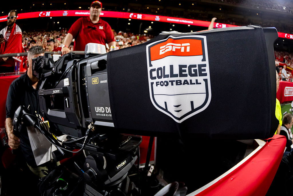 A large ESPN College Football broadcast camera sits on the sideline during a game, with staff and a packed stadium visible behind it.