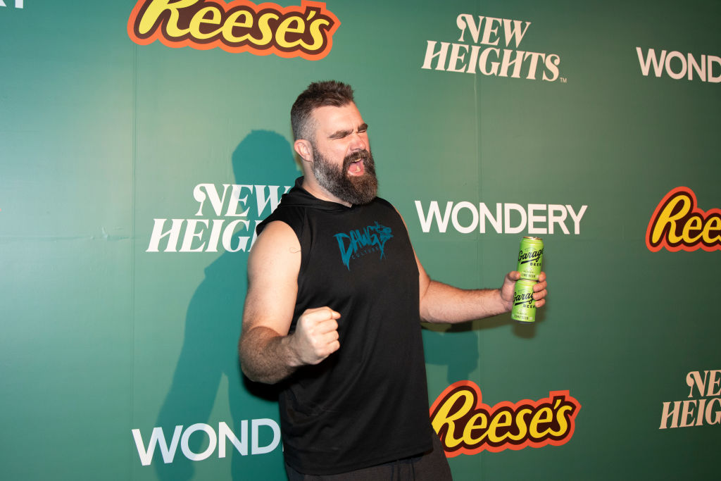 Jason Kelce poses energetically on a green event backdrop featuring Reese’s, Wondery, and New Heights logos, holding a can in one hand with a celebratory expression.