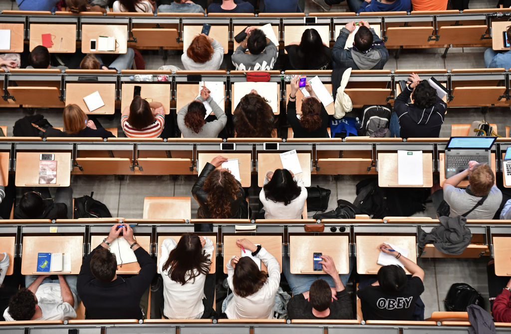 Overhead view of a crowded lecture hall with students seated at long desks, many using notebooks, laptops, or phones.