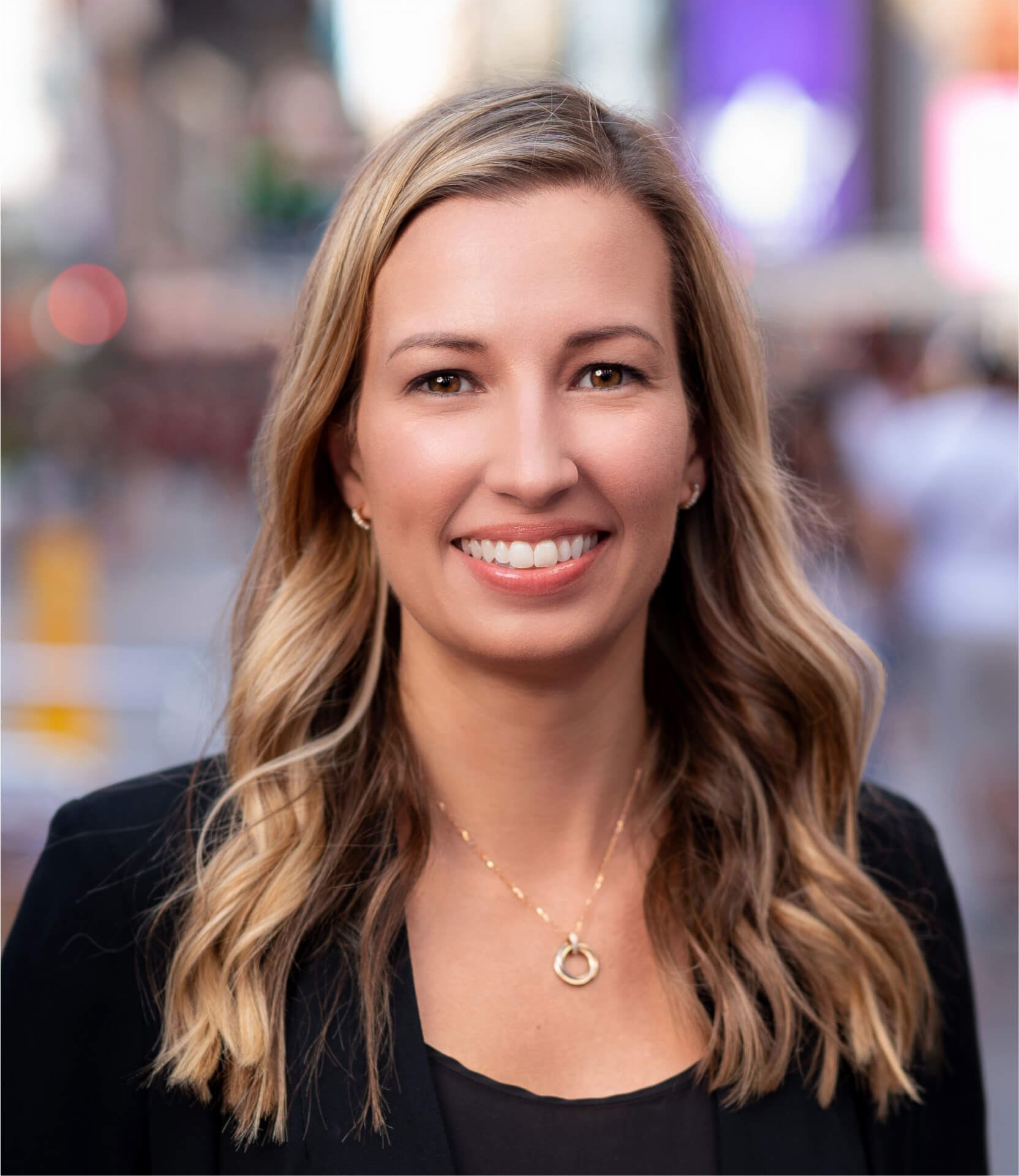 Headshot of a woman smiling, wearing a black blazer and gold necklace, with a blurred city background.