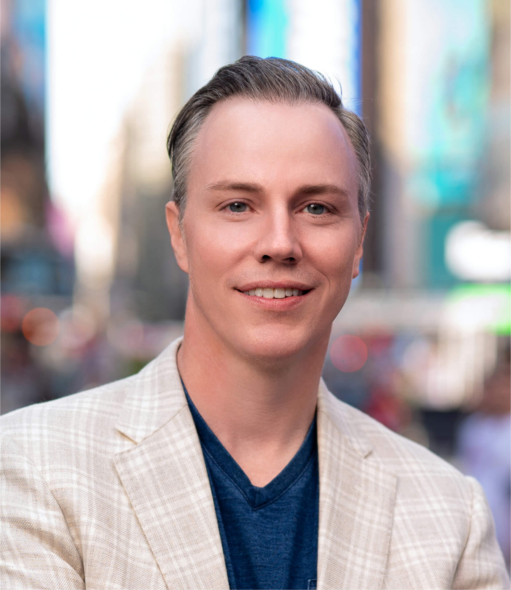 Headshot of a man smiling, wearing a light plaid blazer and blue shirt, with a blurred city background.