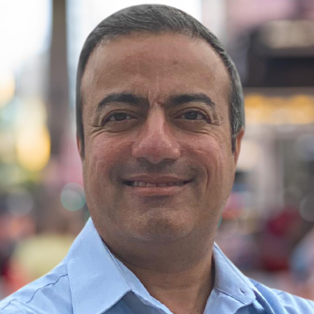 Headshot of a man smiling, wearing a light blue button-down shirt with a blurred city background.
