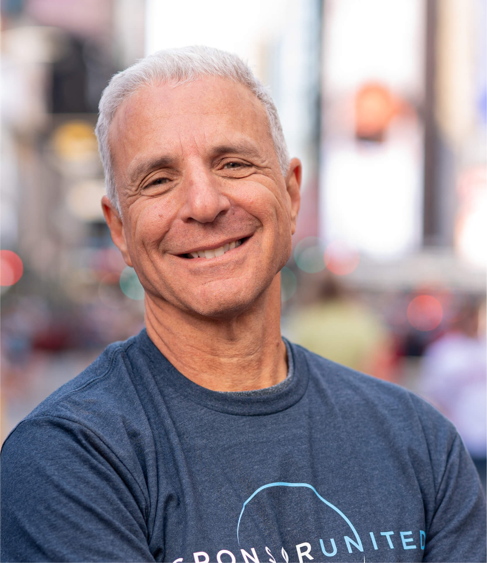 Headshot of a man smiling, wearing a dark T-shirt and a backward cap, with a blurred city background.