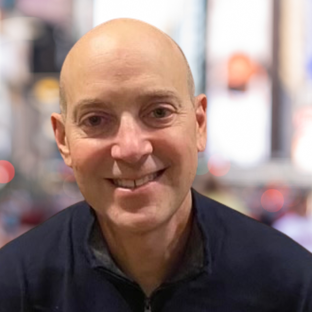 Headshot of a man smiling, wearing a dark shirt, with a blurred city background.