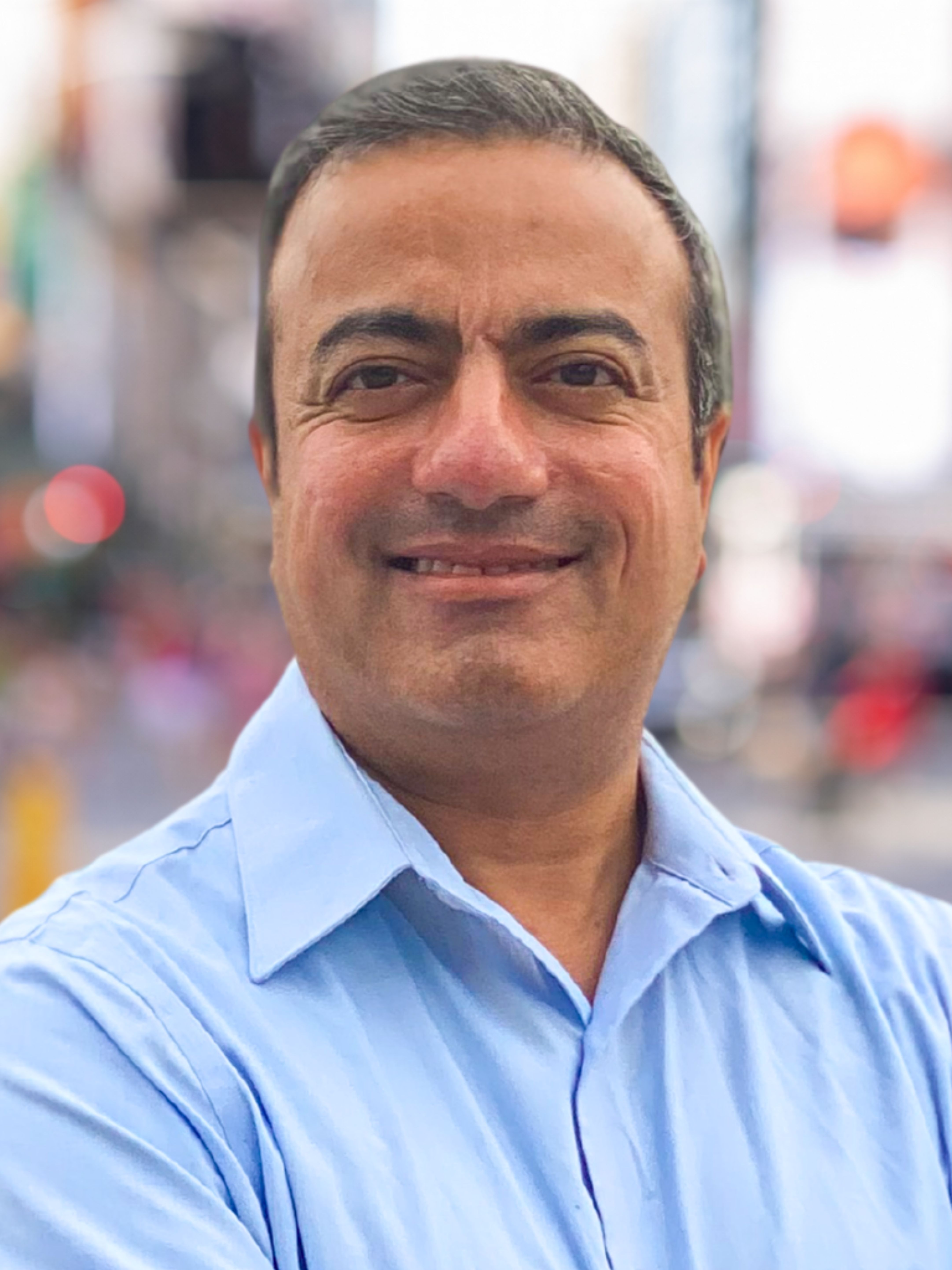 Headshot of a man smiling, wearing a light blue button-down shirt with a blurred city background.