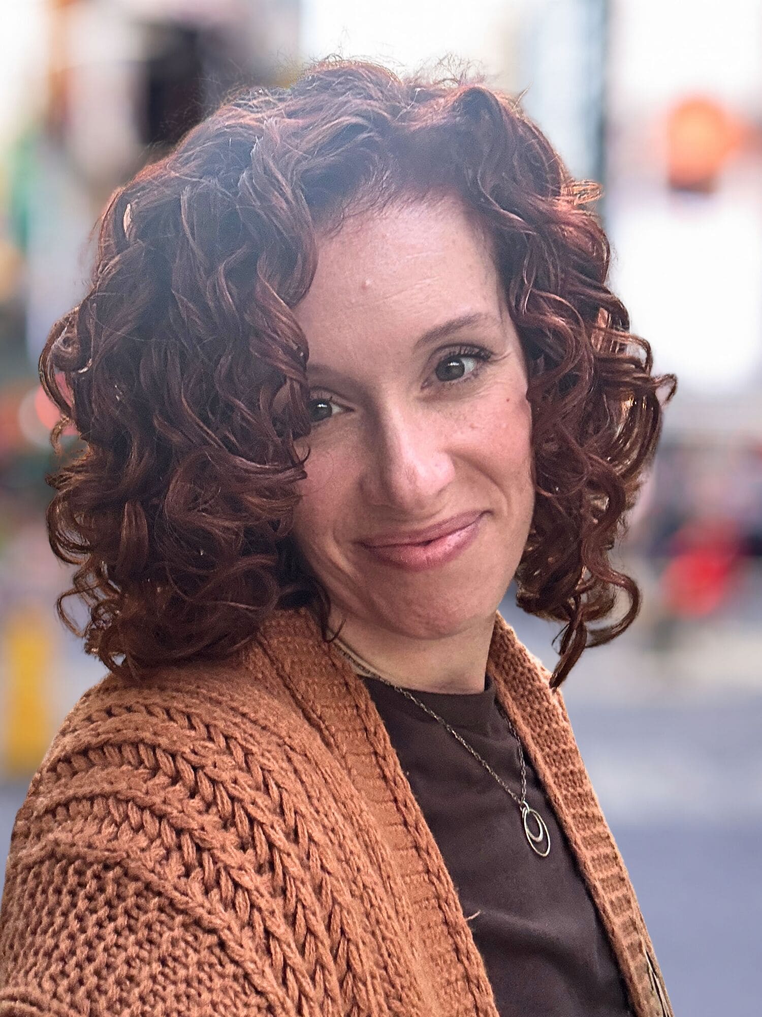 Headshot of a woman smiling, wearing an orange cardigan over a brown shirt with a blurred city background.