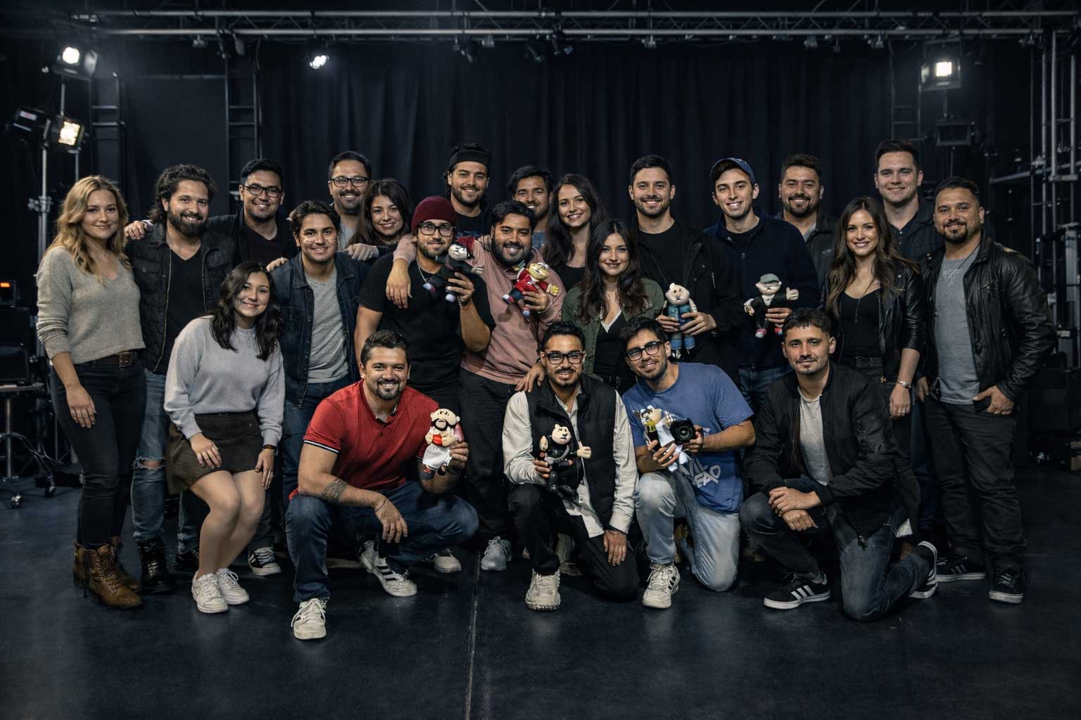 Group photo of twenty-two smiling adults indoors against a black curtain, some holding small cartoon character figurines and a camera.