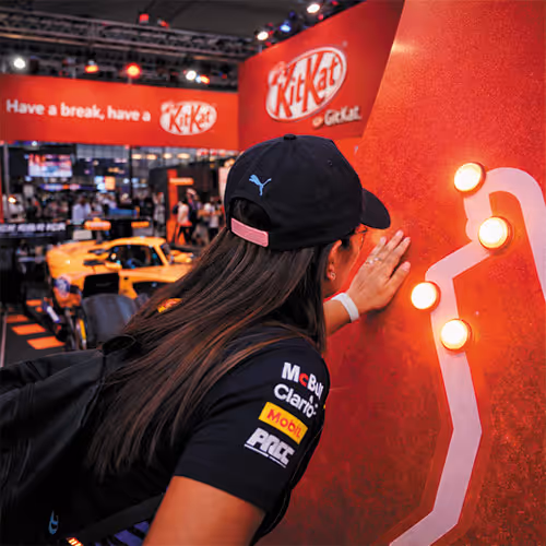 Person wearing a black cap and branded racing shirt touching a glowing wall in a KitKat event space with a yellow race car in the background.