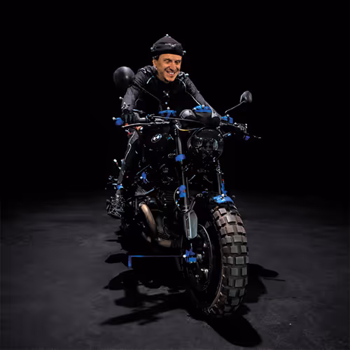 Man wearing motion-capture suit sitting on a black motorcycle with sensors attached, against a dark background.