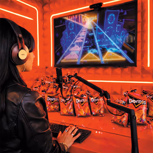 Person wearing headphones playing a rhythm game on a computer surrounded by multiple Doritos bags in an orange-lit room.
