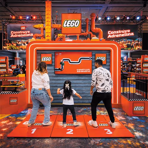 Three people stand on numbered LEGO podiums in front of an interactive LEGO racing game display with orange and black theme.