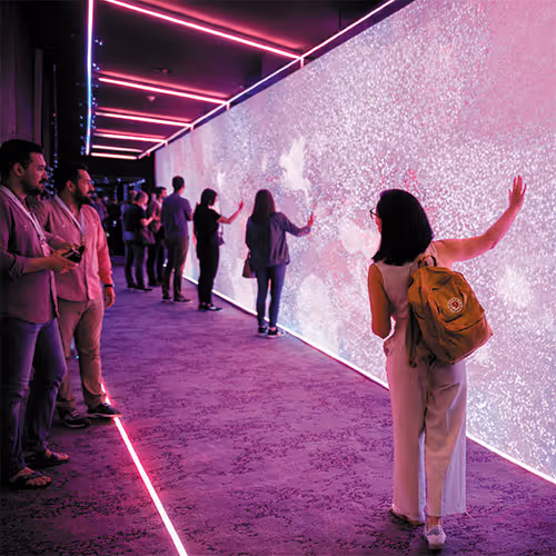 People interacting with a large illuminated digital wall in a dark room lit by pink neon lights.