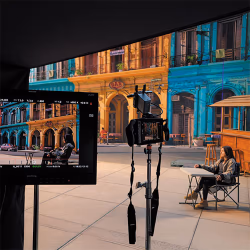 Film camera setup filming a person sitting in a director's chair on a street with colorful colonial buildings in the background.