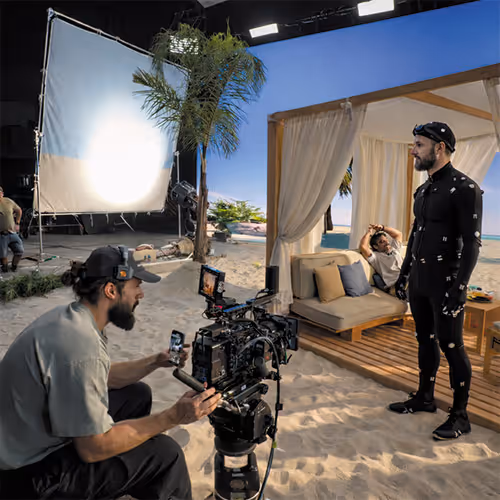 Filmmaker operating camera on a sandy set while an actor in a motion capture suit stands near a beach-themed outdoor seating area with another person reclining on a couch.