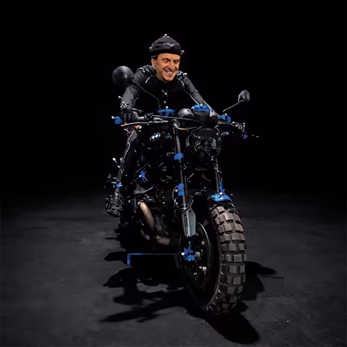 Man wearing motion capture suit and markers sitting on a motorcycle in a dark studio.