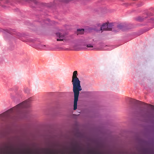 Person standing in a dark room surrounded by immersive pink and purple cosmic or nebula-like projections on the walls and ceiling.