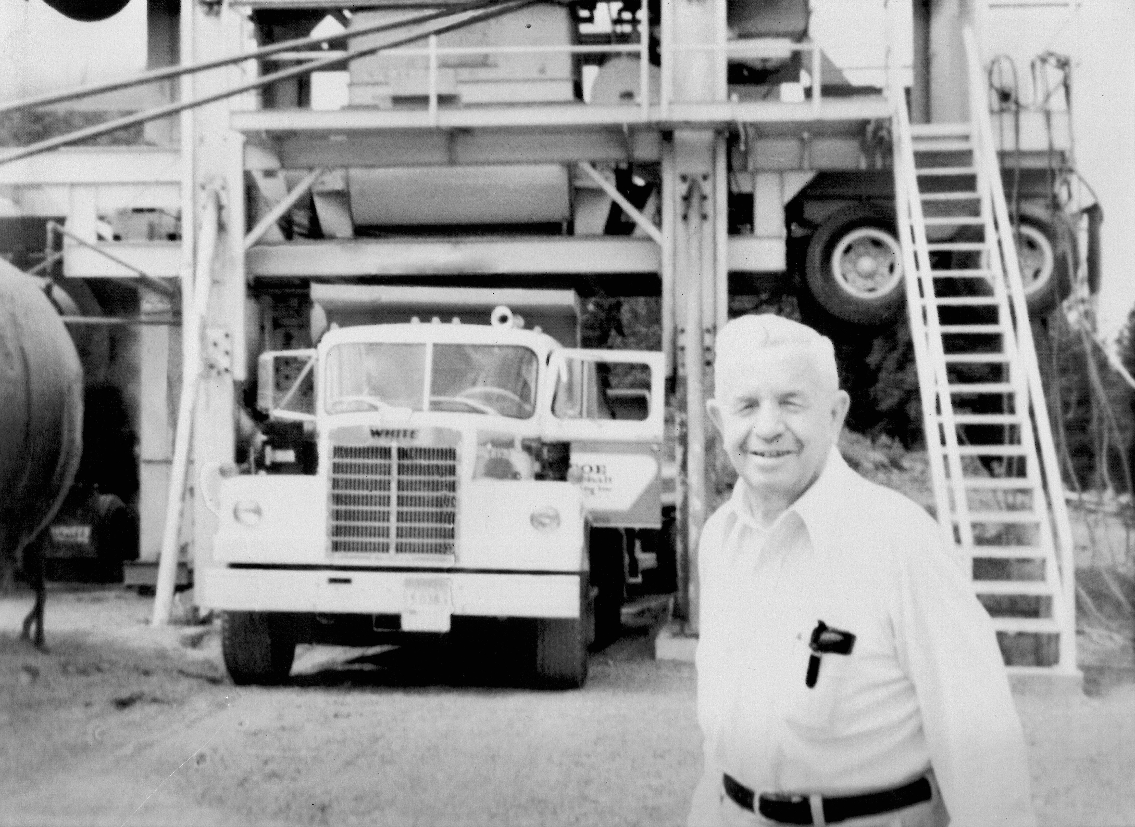 Older man smiling, standing near a large truck and industrial equipment with stairs behind him.