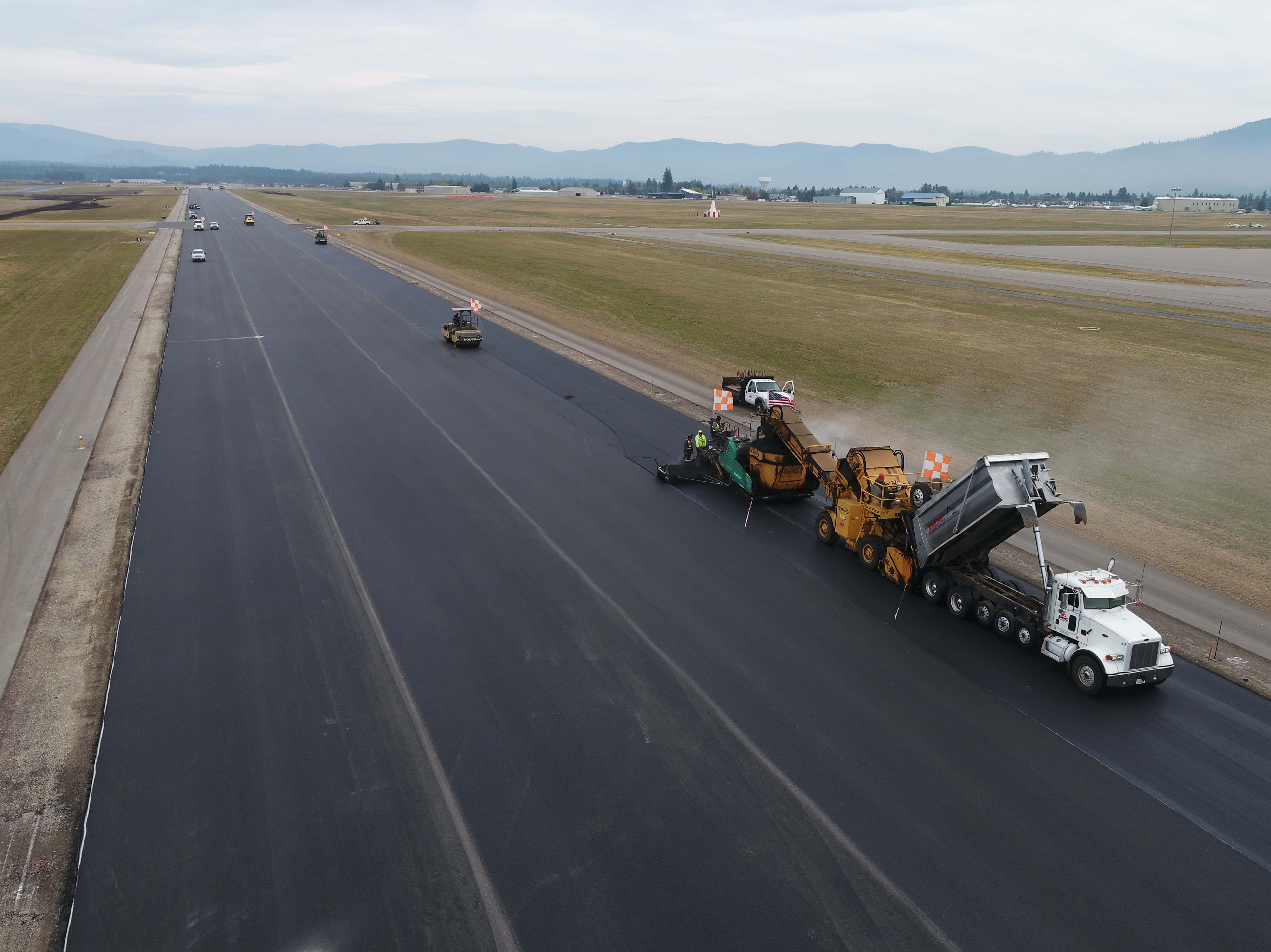 Construction vehicles repaving a long asphalt runway at an airport with hills in the background.