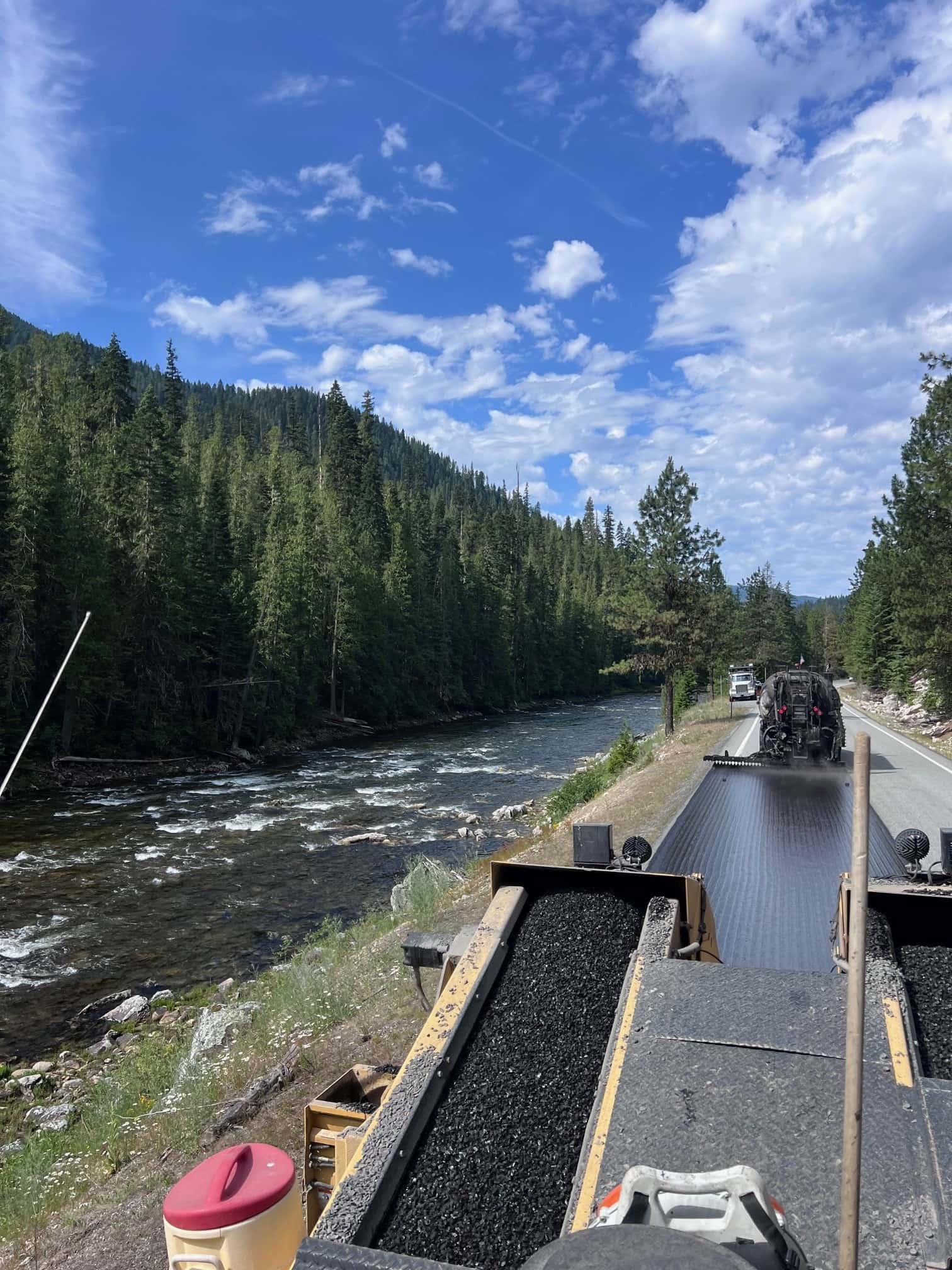 Road construction equipment laying black gravel for chip seal on a mountain road beside a flowing river.