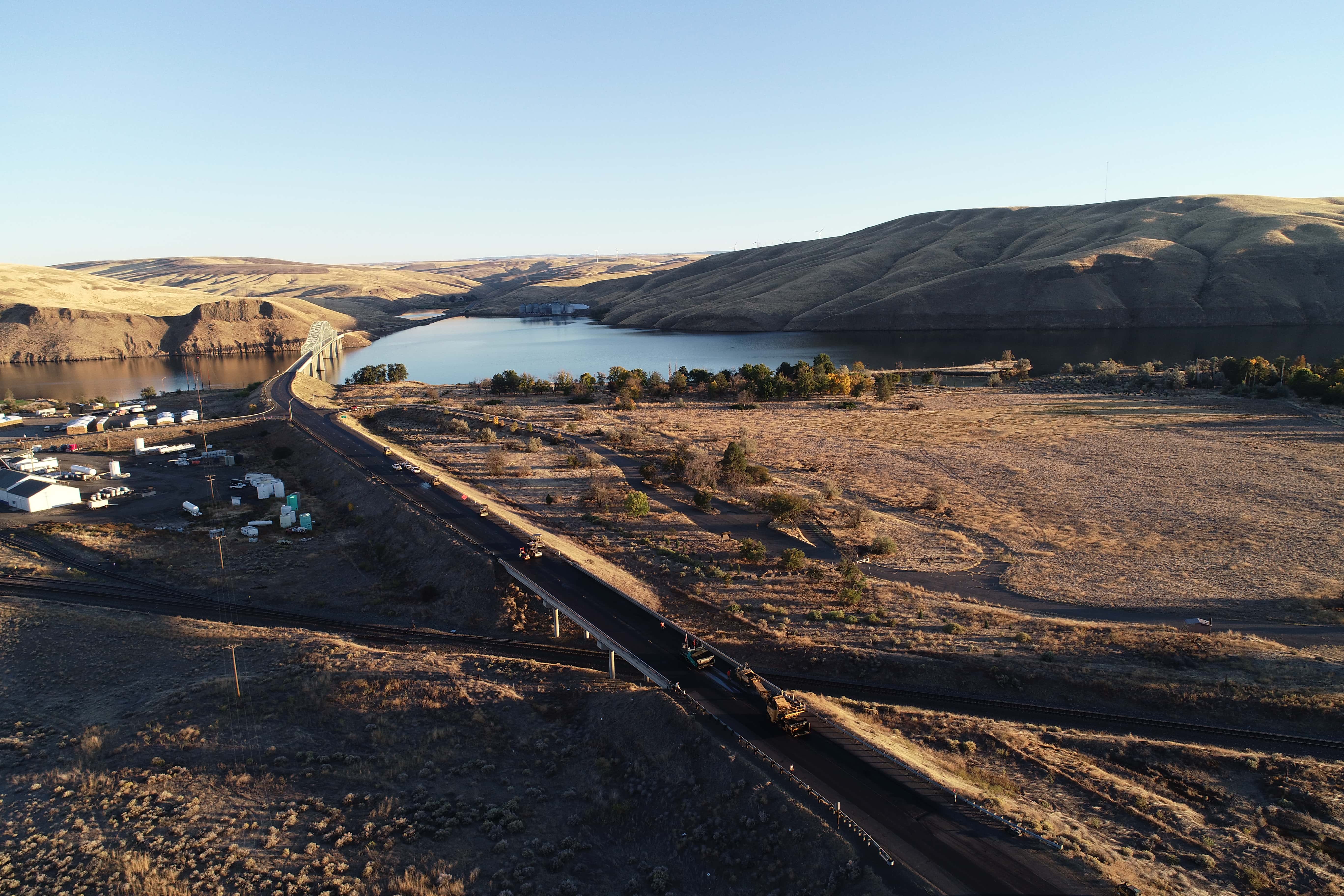 A bridge over calm river waters with dry hills and a road with construction vehicles in the foreground.