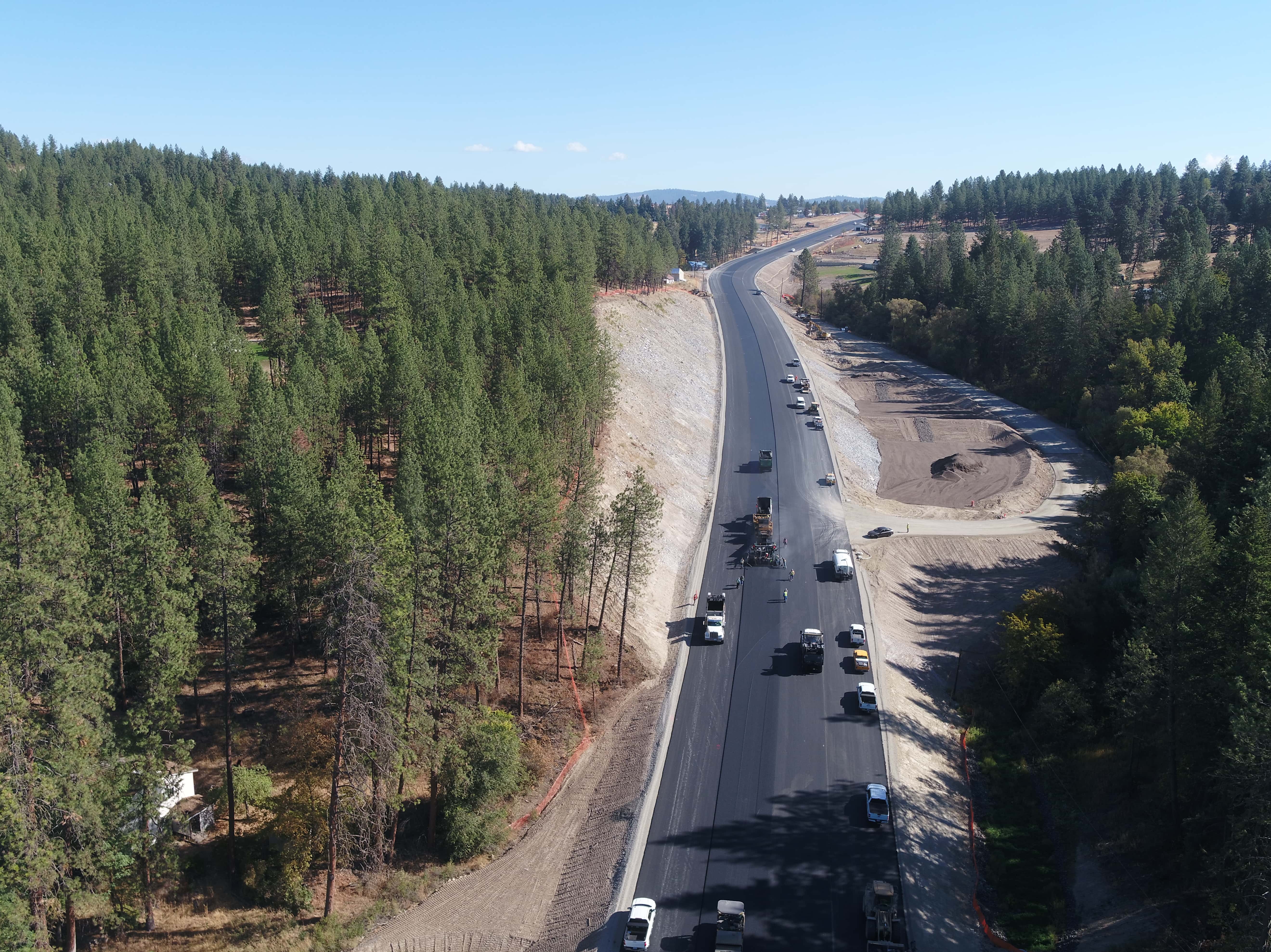 Aerial view of road construction with asphalt paving amid forested landscape on a clear day.