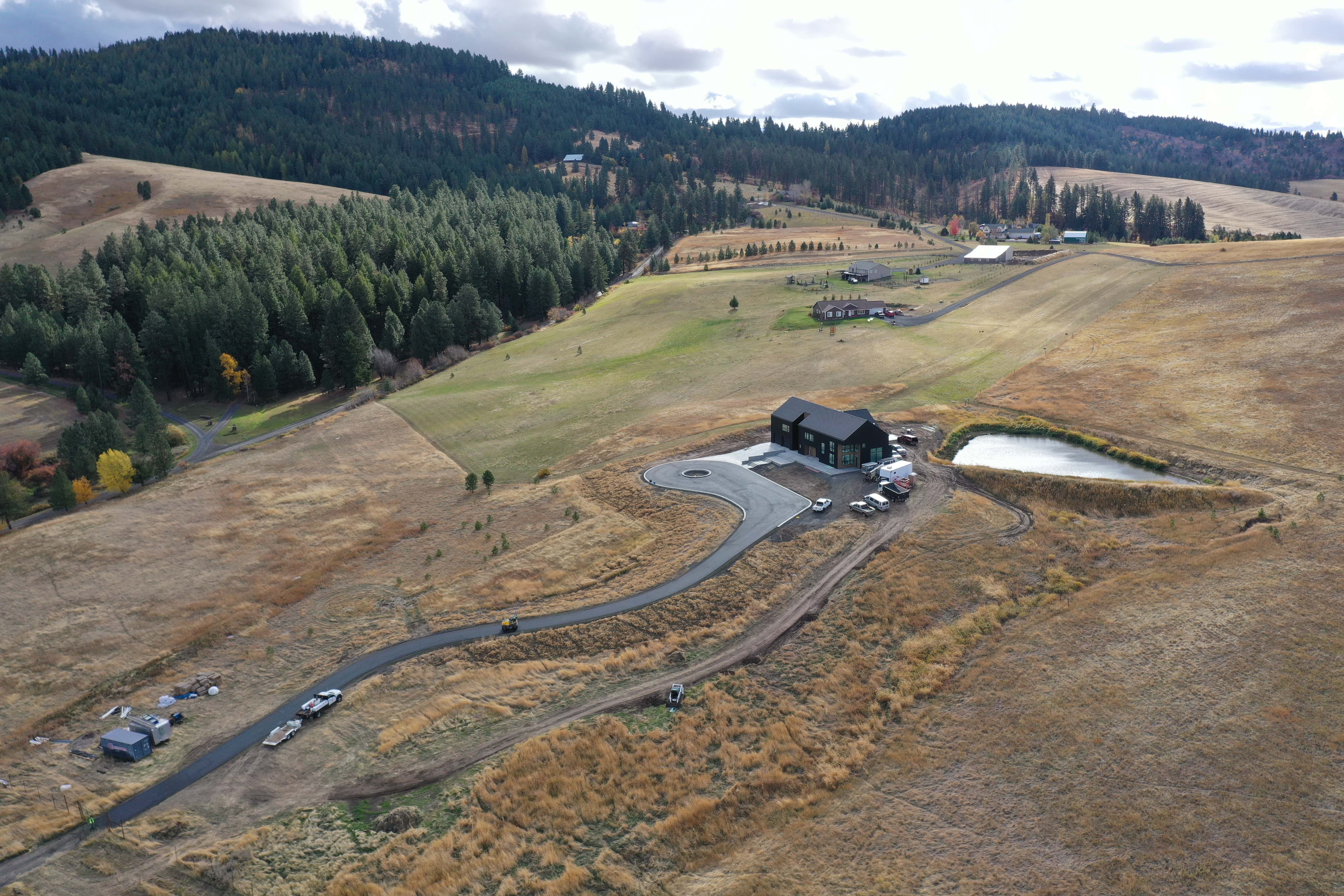 A modern house with a black roof and driveway in a rural grassy area with forested hills and a pond nearby.
