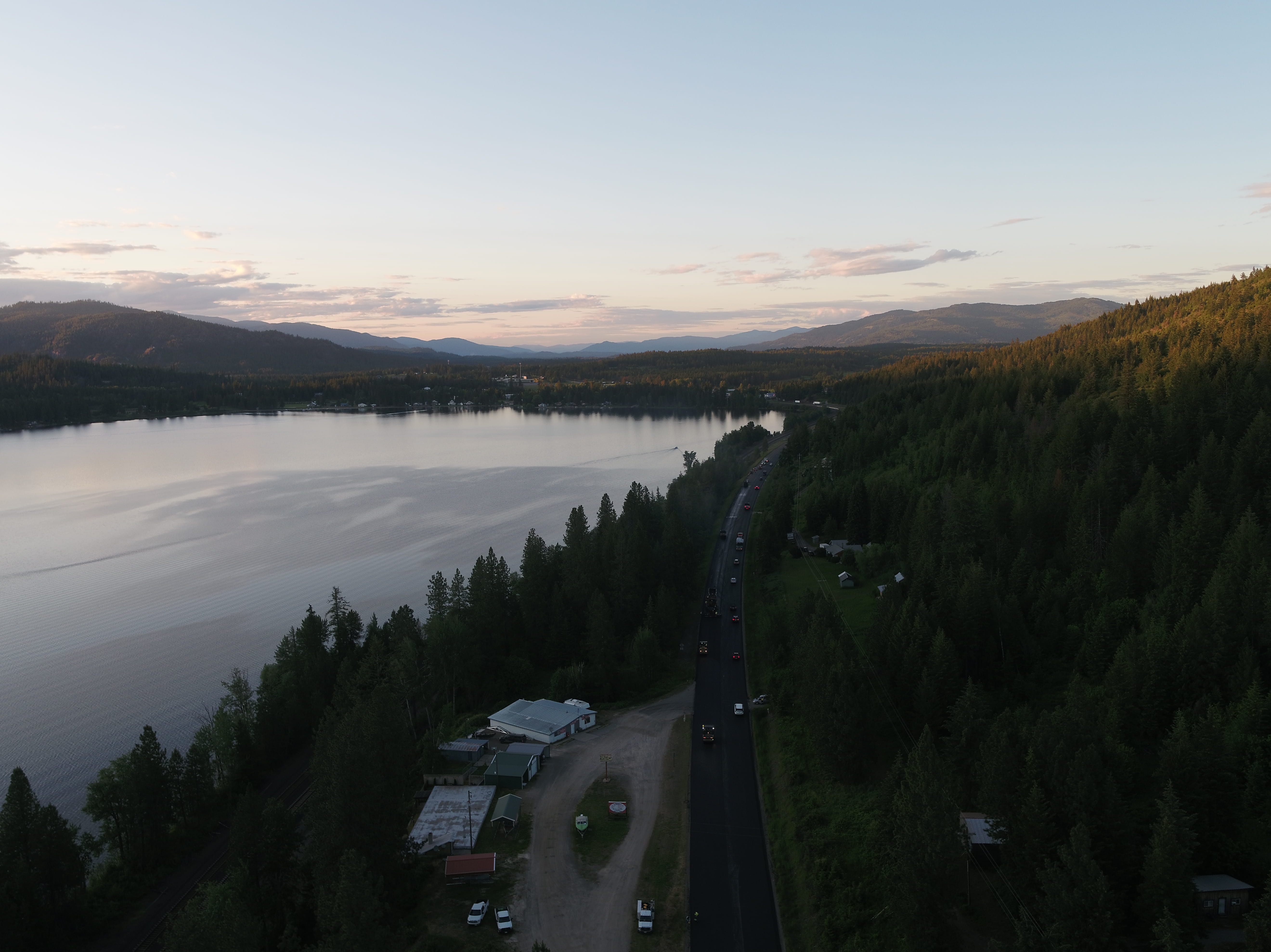 A road runs alongside a large lake surrounded by dense forest and mountains under a clear sky at dusk.