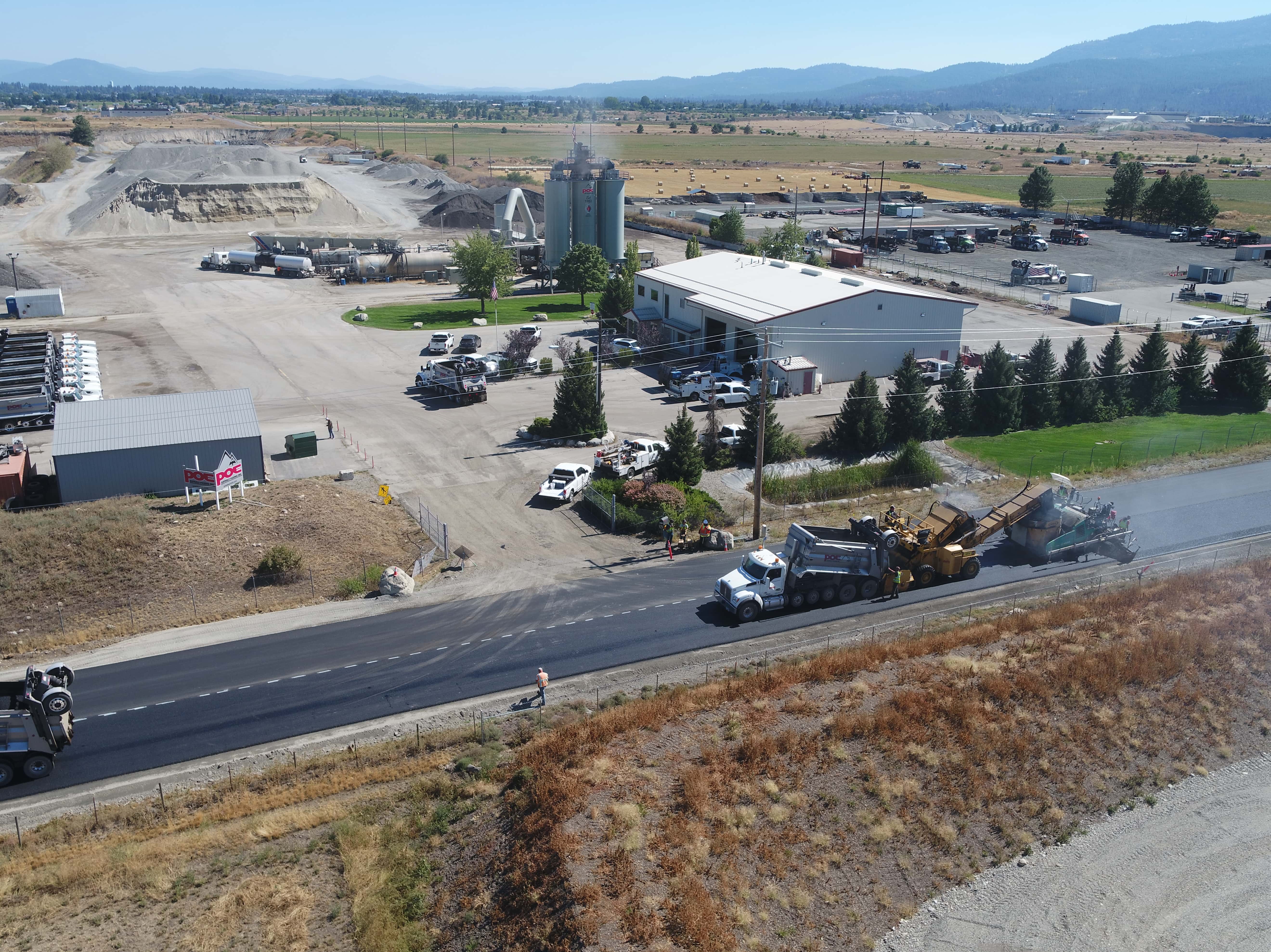 Workers and trucks paving a road near industrial buildings and fields with mountains in the background.