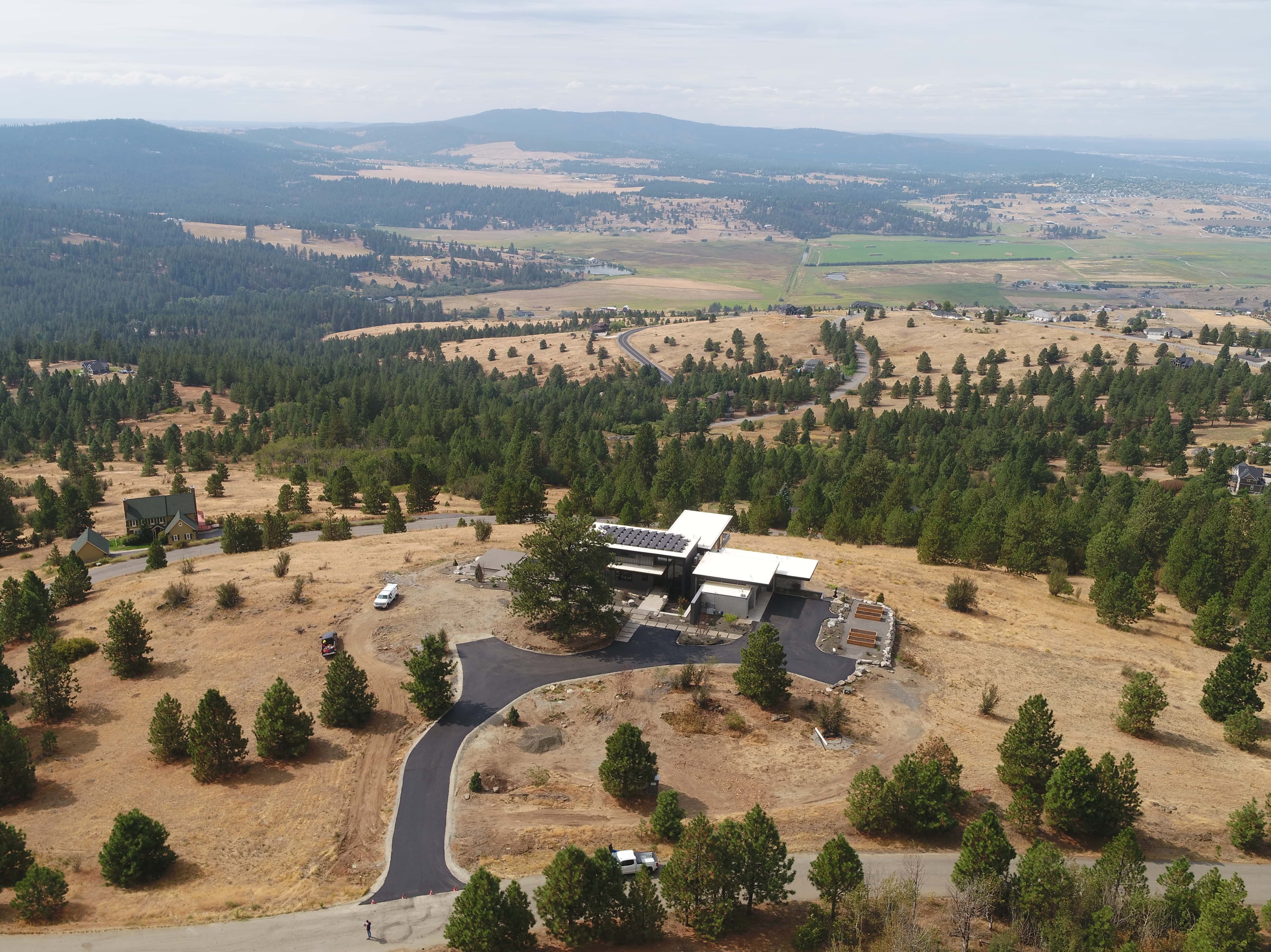 Modern house with solar panels on roof surrounded by dry landscape and scattered evergreen trees.