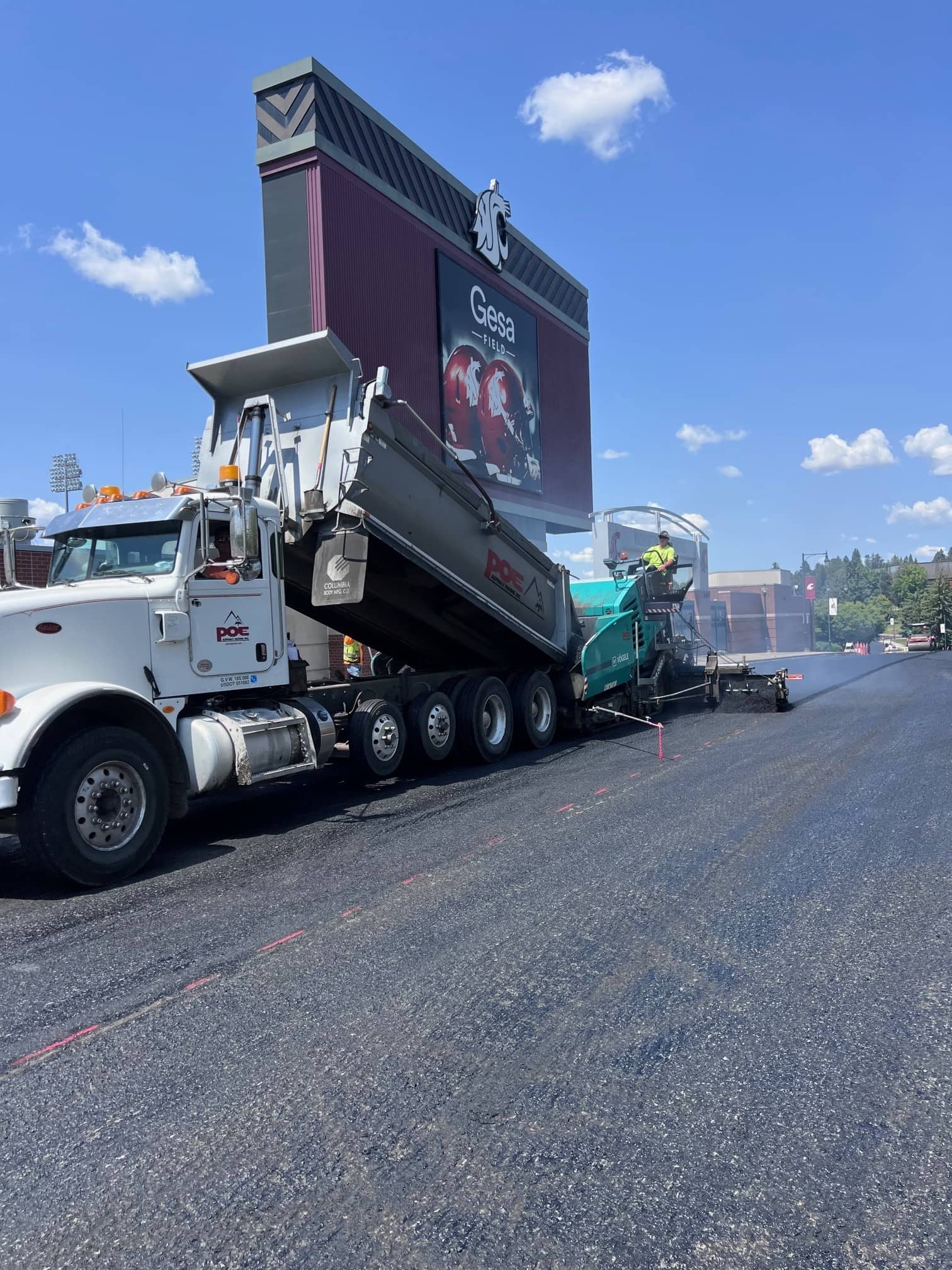 Construction truck and paver laying asphalt near Gesa Field stadium scoreboard under blue sky.