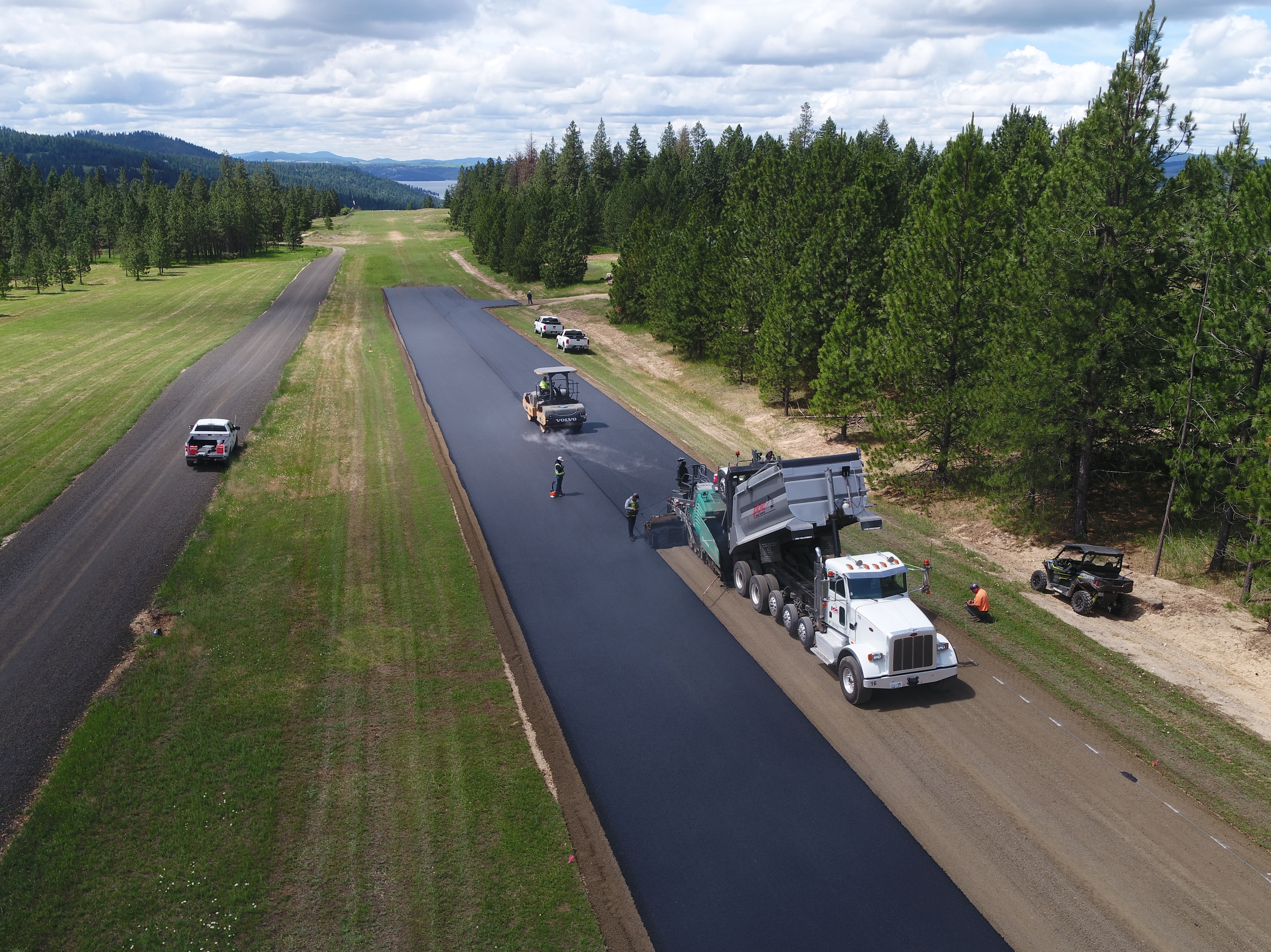 Construction workers paving a long narrow road surrounded by grass and trees under a cloudy sky.