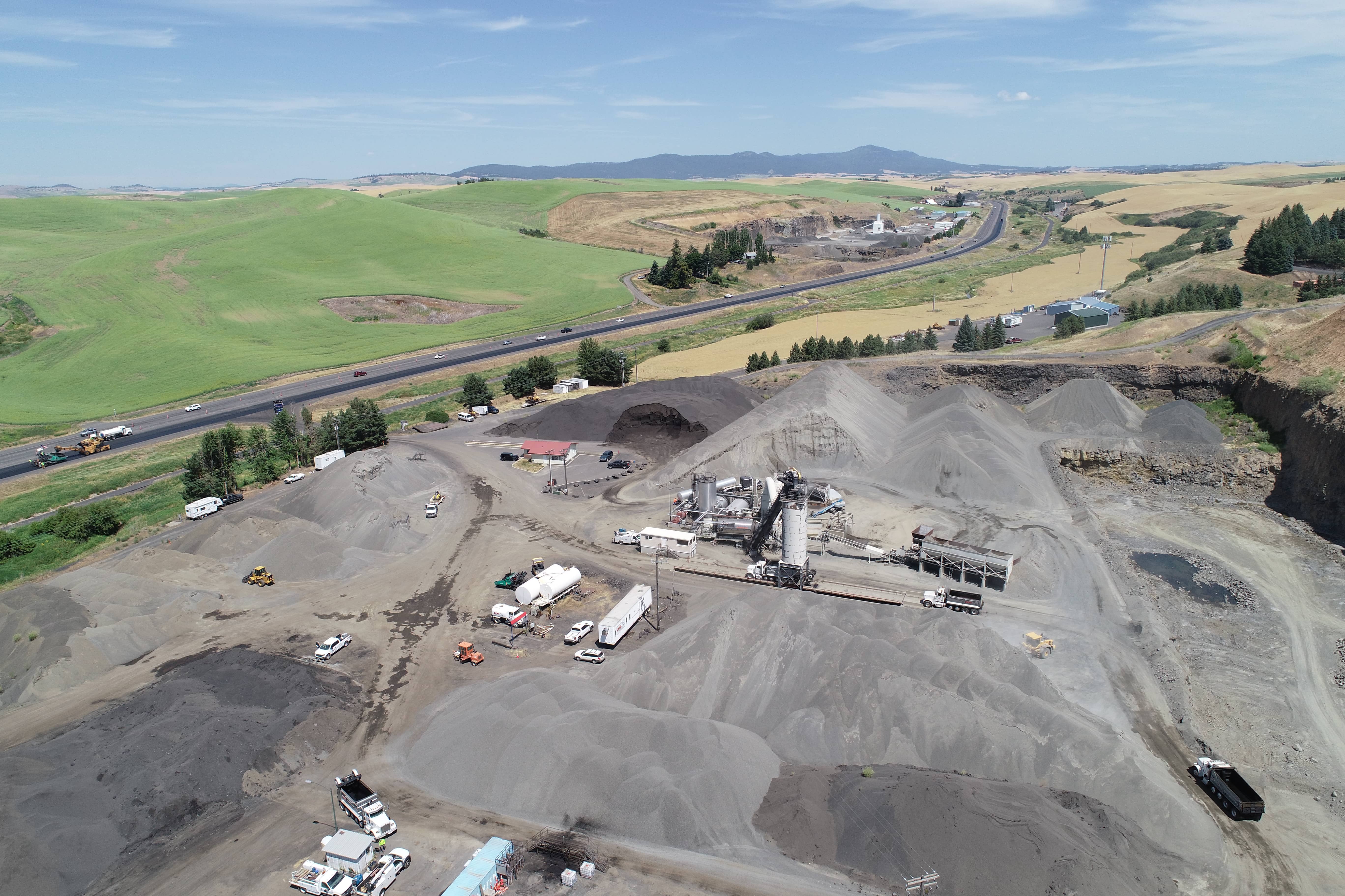 Aerial view of an asphalt plant with large gravel piles and trucks near a highway and green fields.