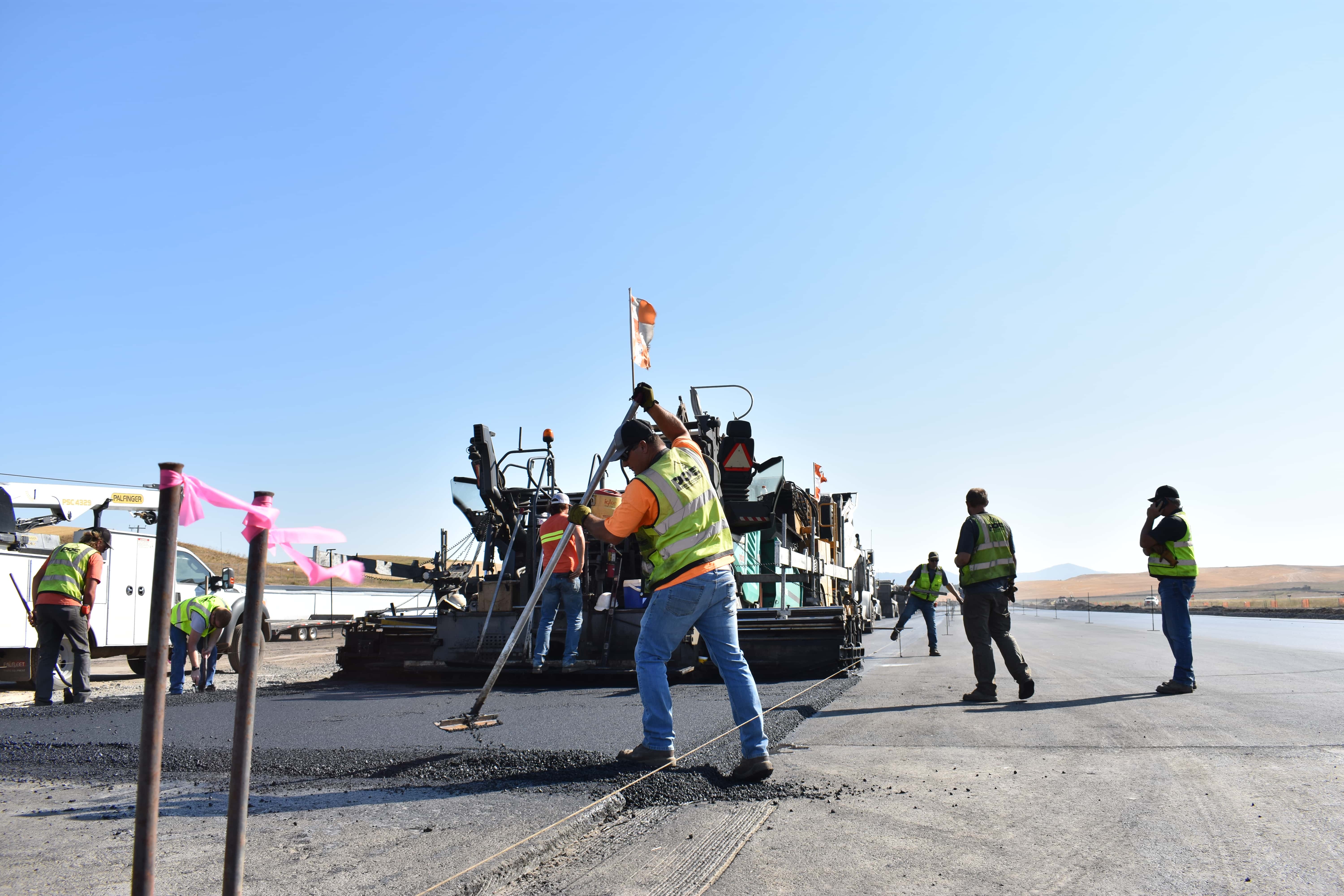 Workers wearing safety vests paving and smoothing asphalt on a road under clear blue sky.