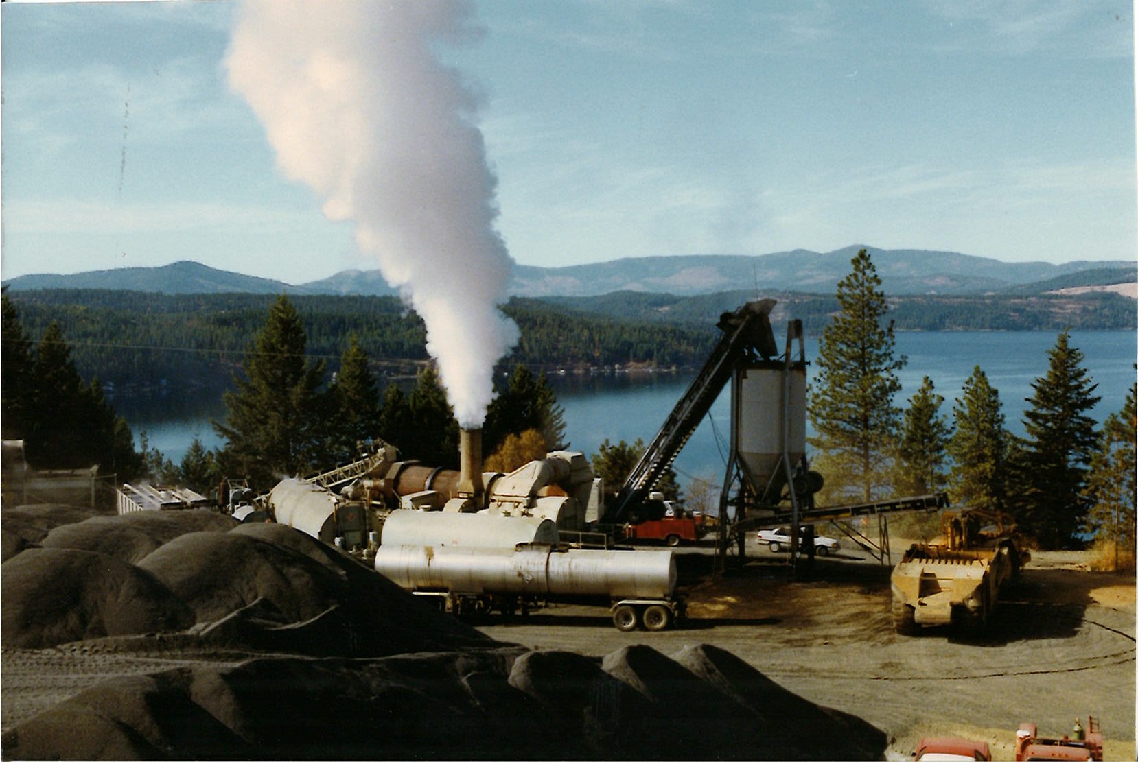 Industrial site with steaming machinery near piles of gravel and a lake surrounded by forested hills.