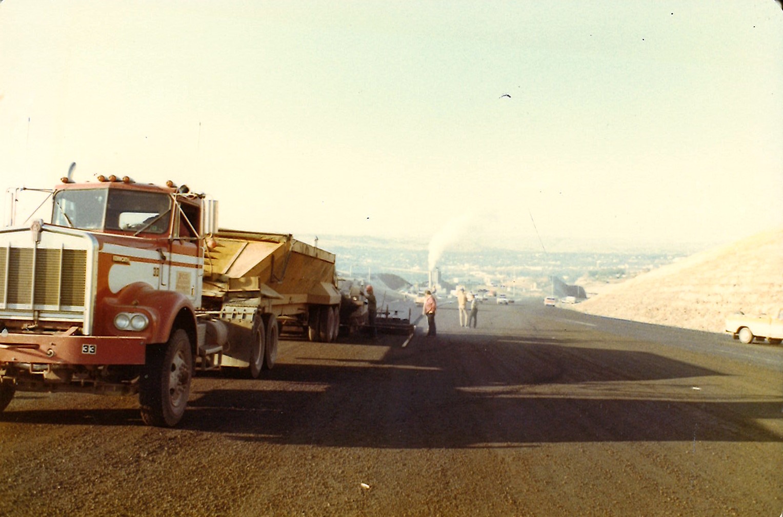 Large red and white Kenworth truck with trailer on a newly paved road, workers and distant city view.