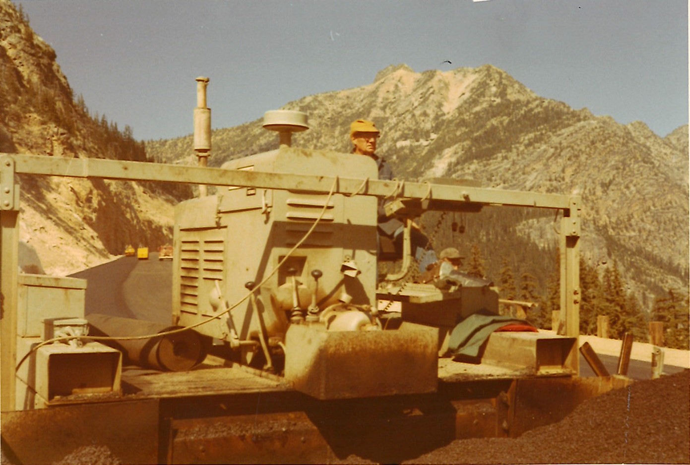 Construction worker operating heavy machinery on mountain road with rugged mountain peaks behind.