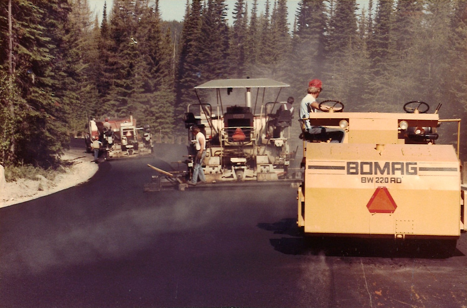 Road construction machinery paving a forested road with workers operating equipment.