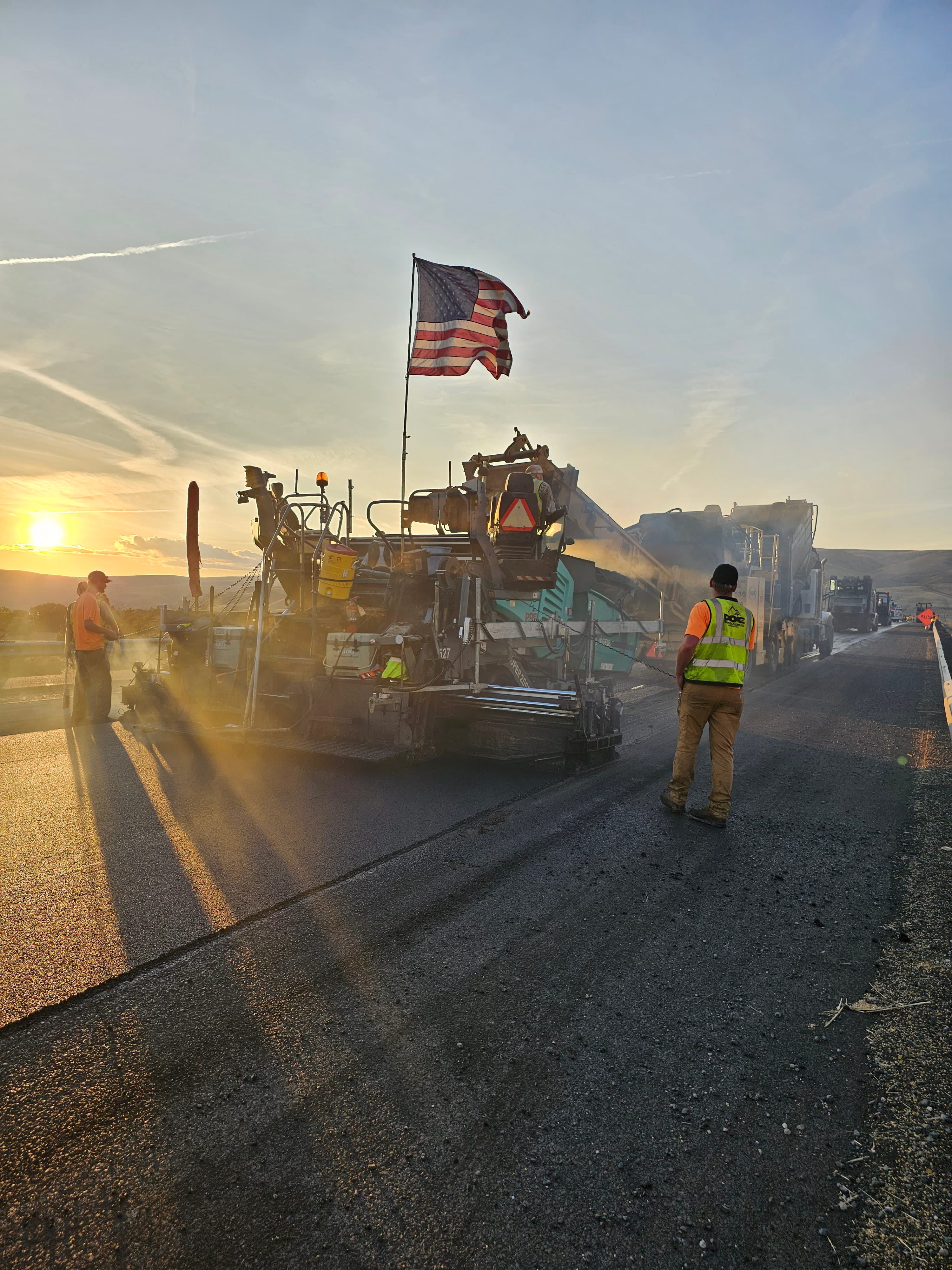 Workers in orange vests paving a road at sunset with an American flag on the machine.